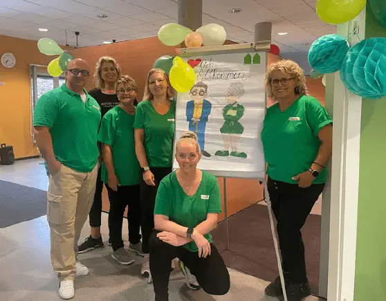 Team der Nachbarschaftshilfe steht gemeinsam vor einem Banner und lächelt in die Kamera.
