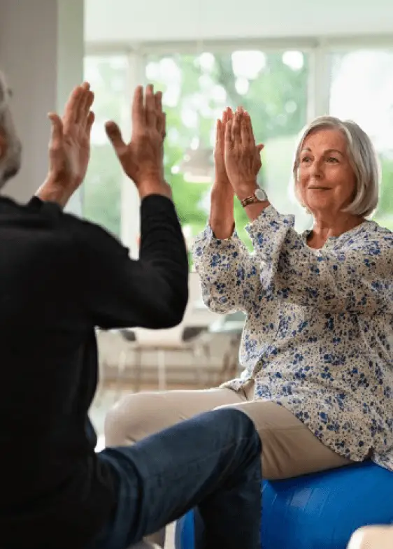 Seniorin hebt lächelnd die Hände und nimmt an einer Bewegungsübung teil.
