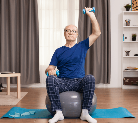 Senior sitzt lächelnd auf einem Stuhl und hebt den Arm während einer Bewegungsübung.